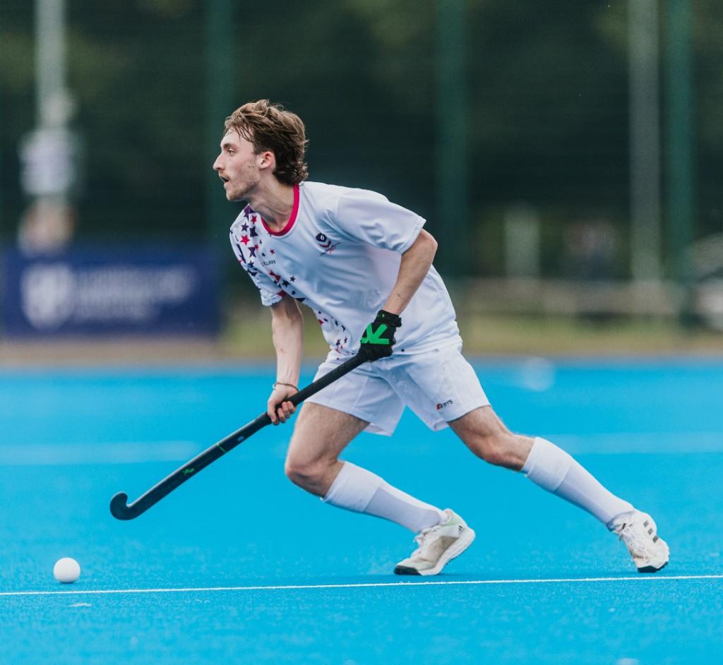 Dominic Hubble playing hockey on a blue pitch in Loughborough kit
